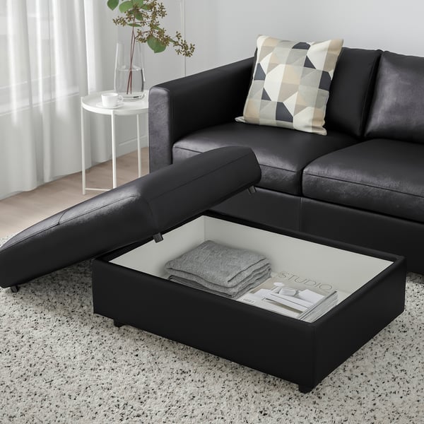 Black leather sofa with open storage footstool, grey pillow, white side table, and rug in living room.