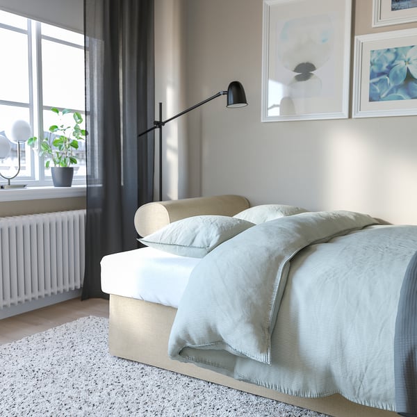 Modern bedroom with grey curtains, bed made with light blue blanket, green pillow, art on wall, plant by window, white radiator, lamp near bed.