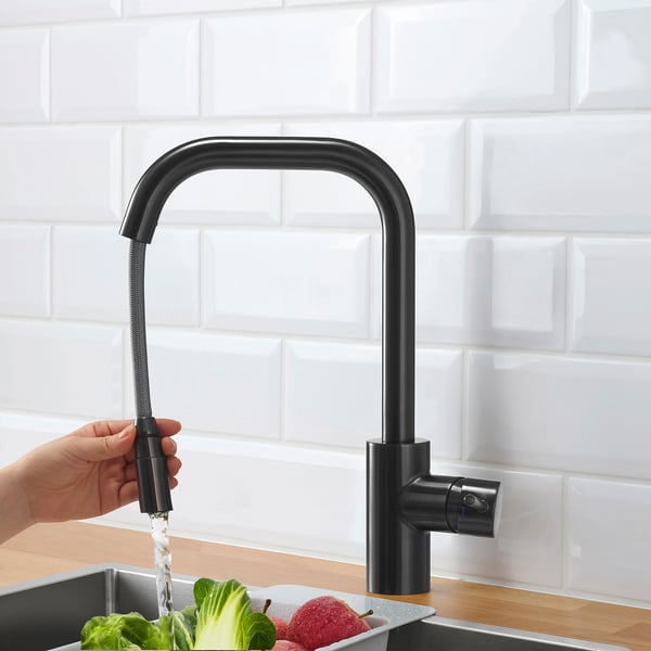 Hand holding black tap spraying water into sink with fruits and vegetables.