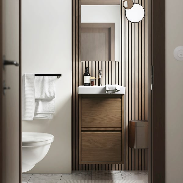 Modern bathroom ÄNGSJÖN cabinet in brown oak, wall-mounted, with sleek minimalist design and vertical handles, smaller than nearby toilet