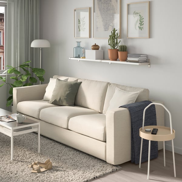 Modern living room with beige sofa, wooden floor, grey curtains, wall art, and plants. White shelf with books and plants.