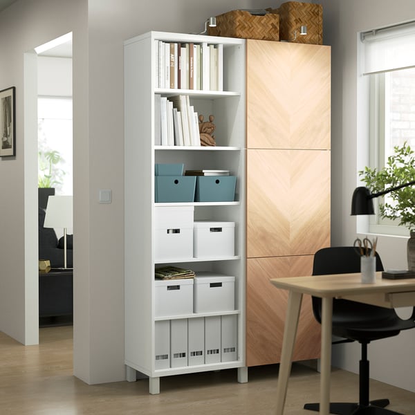 BESTÅ white tall storage unit with open/closed compartments, holding books and decor, surrounded by wood furniture in a bright room.