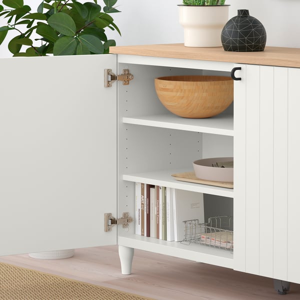 Open white cabinet displaying bowls, plates, books, with gold hinges. Plant and decorative items on top.