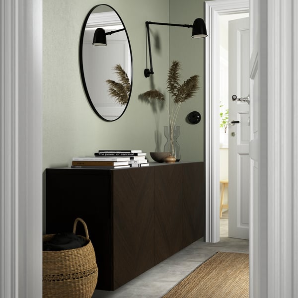 Modern entryway with BESTÅ sideboard, woven basket, light green wall, mirror, black sconce, dried plants in vase, and stack of books.