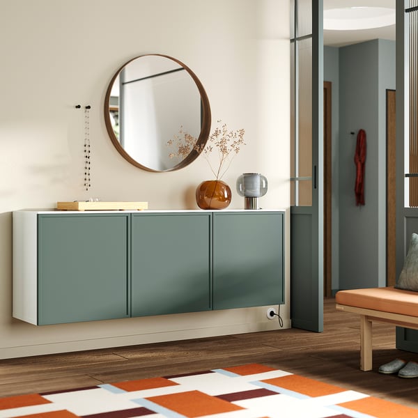 Modern entryway BESTÅ sideboard in green with glossy white top. Round mirror above, decorative vase and lights add style. Wooden bench nearby.