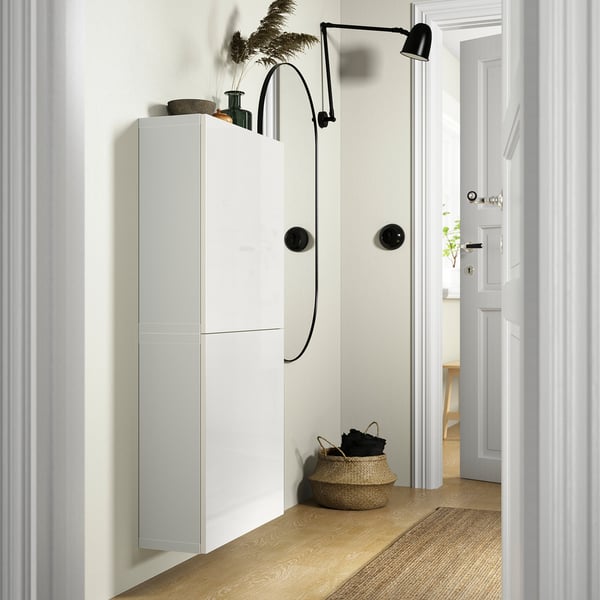 White BESTÅ cabinet, narrow design, wall-mounted with black knobs, beside a tall plant and basket, near white door.