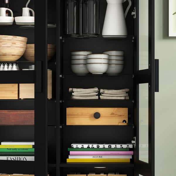Black display cabinet with glass doors, showing stacked bowls, vases, folded towels, wooden tray, and colourful books on shelves.