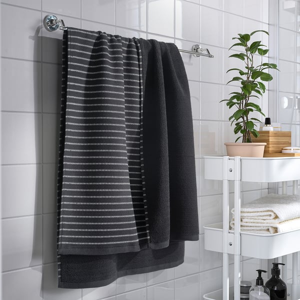 A modern bathroom with a grey towel on a chrome bar, next to a shelf holding folded towels, a plant, and toiletries.