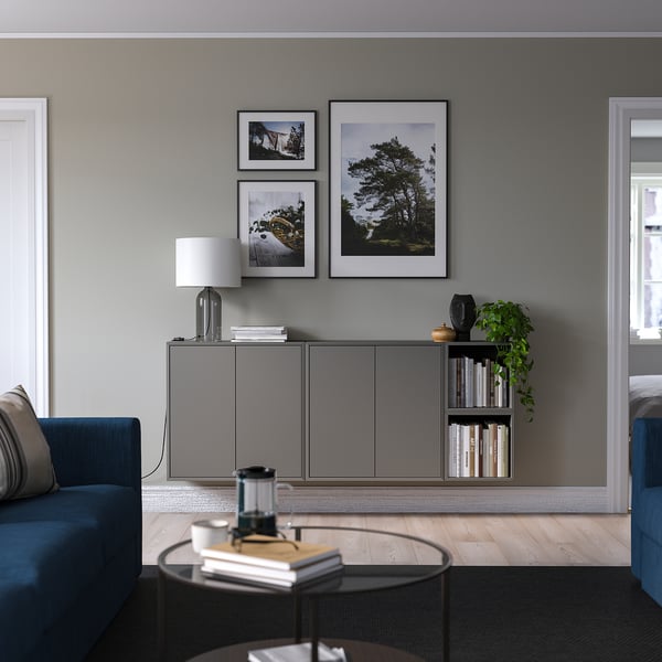 Modern living room with blue sofa, round table, and grey EKET storage. Wall decor includes framed photos and a plant.