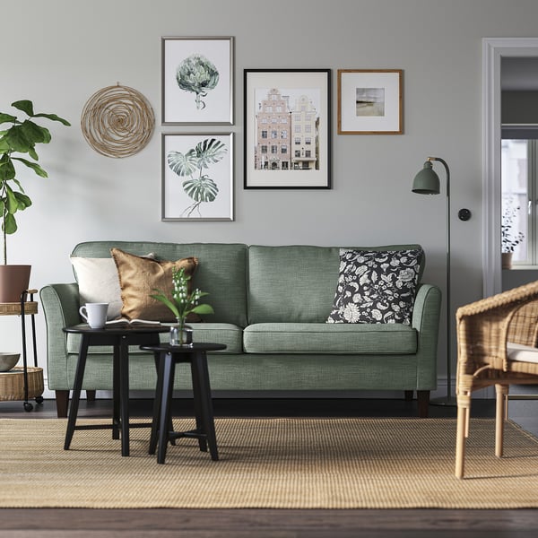 Modern living room with EKHOLMA sofa, green, plush, 3-seater. Wall art, floor lamp, wicker chair, and rug. Clean, minimalist design.