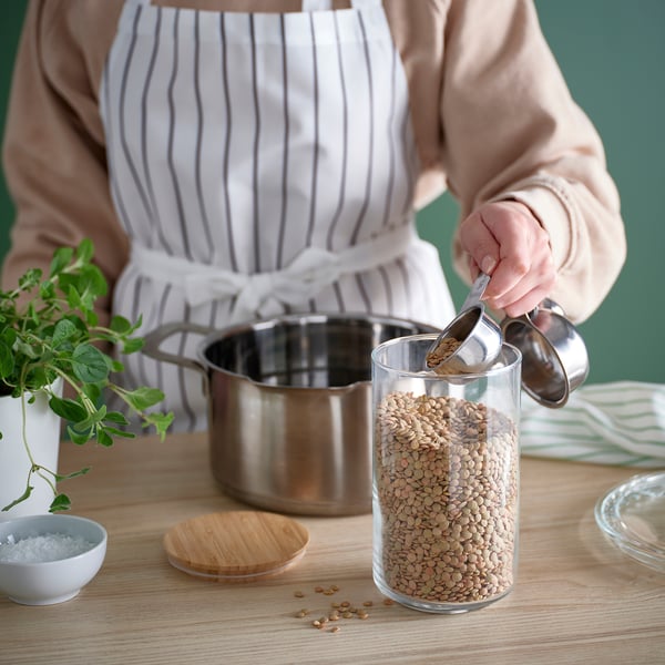 Person measuring green lentils with a stainless-steel scoop from a transparent glass jar for cooking.