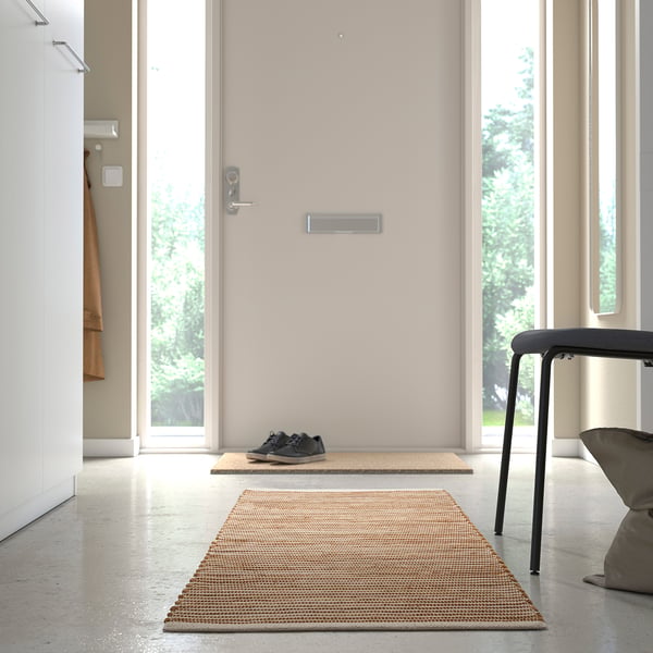 A minimalist hallway with a beige doormat, black trainers, and a grey chair. A handwoven rug decorates the polished floor.