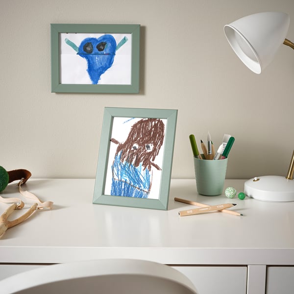 Two childs drawings in light blue frames: one yoda, one person. Frames sit on white desk with art supplies.