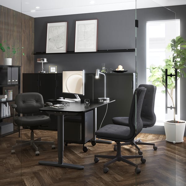 Modern office black desks with ergonomic chairs, surrounded by dark wood accents and decorative plants. Two framed city maps adorn the wall.