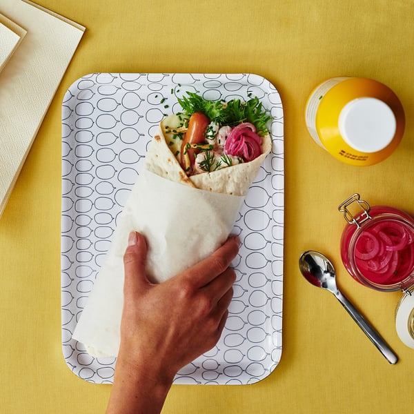 A person holding a wrap with vegetables on a patterned tray next to a jar of pickles and a yellow cup.