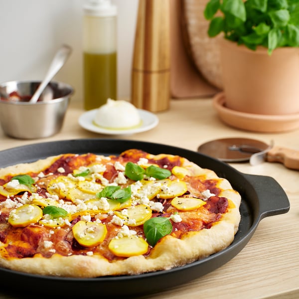 Black cast iron HAVSTULPAN pizza pan with homemade pizza, veggies, and feta on wooden table.