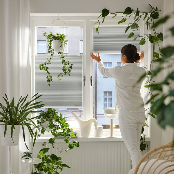 A person adjusts pleated blinds on a window, surrounded by hanging and potted plants. The blinds block out light effectively.