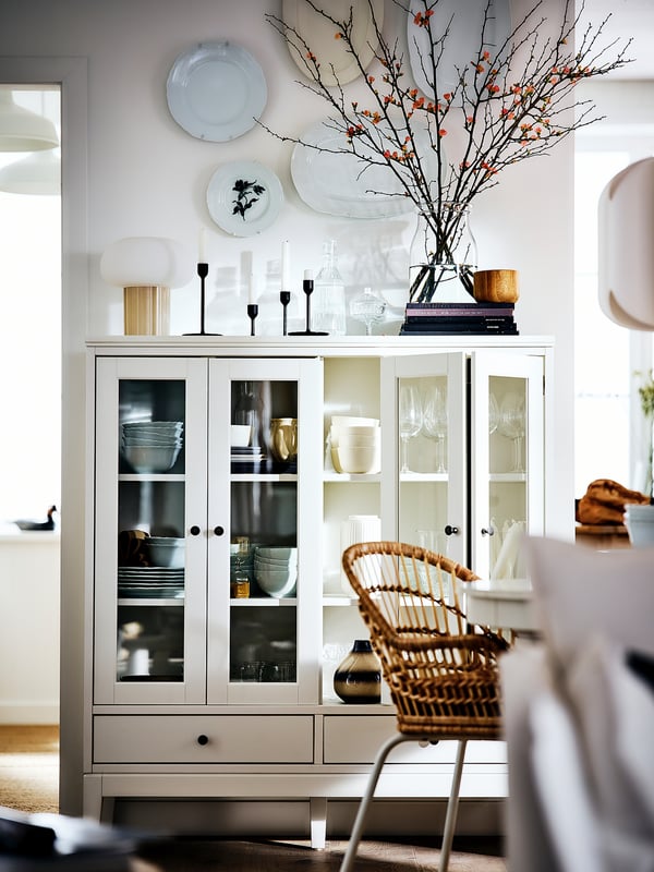 White IDANÄS cabinet with glass doors, holding dishes and vases, topped with decorative plates, candles, and floral branches.