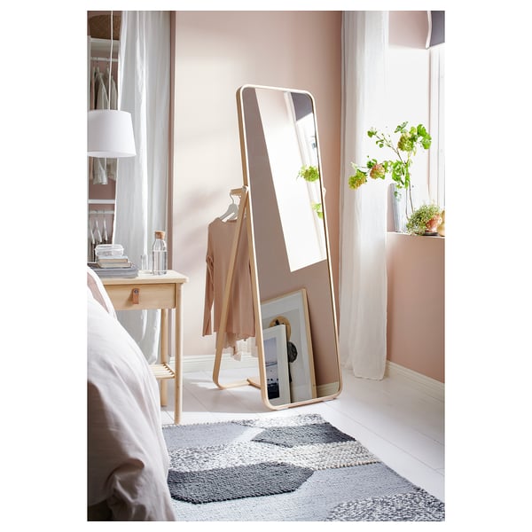 Bedroom with white bed, wooden table, and gold-framed floor mirror reflecting clothes rack and window.