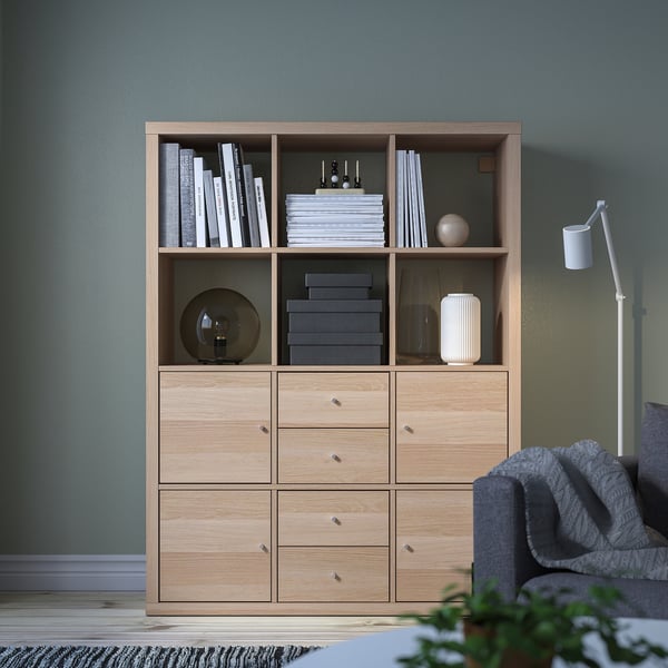 KALLAX light oak bookcase with drawers, hosting books, decorative items, and a lamp, fits well in a cosy green room with a grey couch nearby.