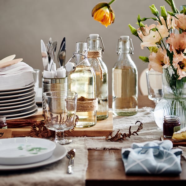 Wooden table set for meal, featuring KORKEN jars with beverages, plates, cutlery, flowers, and cloth napkins.