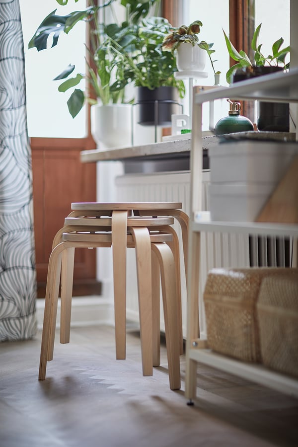 Modern, wooden stools stacked, placed near a window with green plants, brightening cosy space.