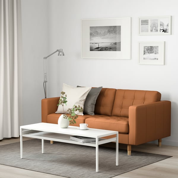 Modern living room with brown sofa, pillows, coffee table with items against white wall with photos.