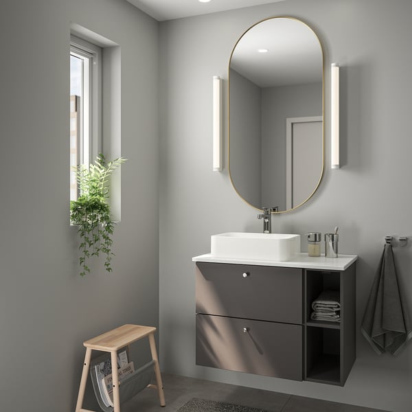 Modern bathroom with large gold-framed mirror, dark grey floating vanity, wooden stool, plant, and grey towels.