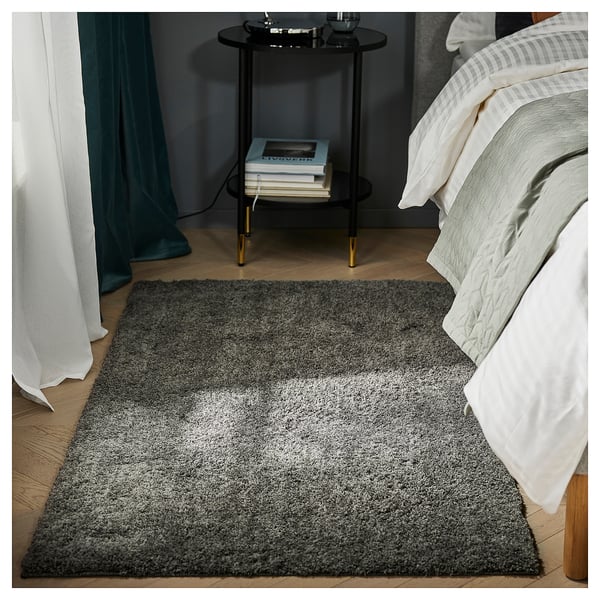 Bedroom with grey LINDKNUD rug, white bedding, black bedside table, teal curtains, and stack of books.