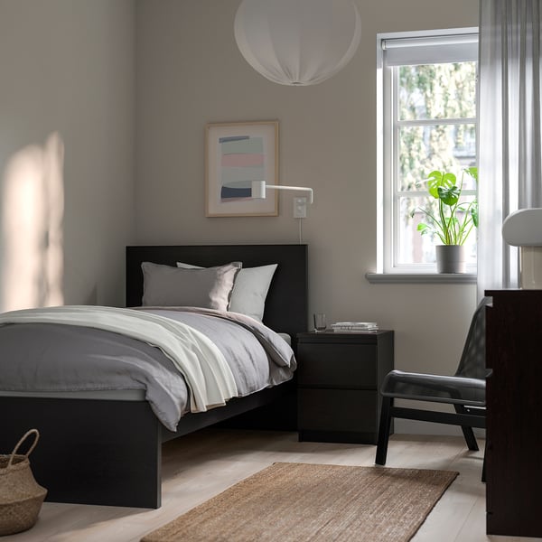 Modern bedroom with grey bed, white linens, black bedside table, lamp, large window, plant, and rug.