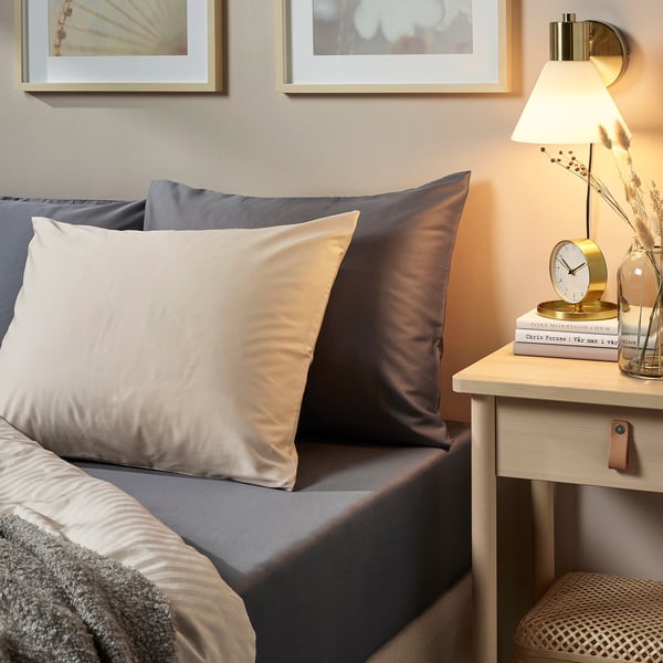 Beige pillows on grey bed with bedside lamp, clock, books, and dried flowers.