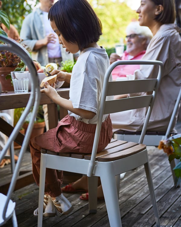 Outdoor dining scene. Child in grey shirt sits at white and wooden NORRMANSÖ table. Adults talk nearby.