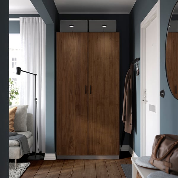 Modern bedroom featuring tall wooden PAX wardrobe, larger than the grey chair and plant nearby. Beige coat hangs on hooks beside a white door.
