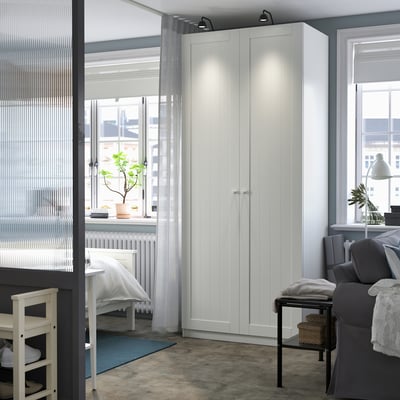 Modern, minimalist workspace featuring a tall, sleek white PAX wardrobe, grey curtains, and cosy seating. Clean, simple design.