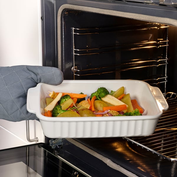 Person placing rectangular baking dish with vegetables into oven, using handle for easy grip.