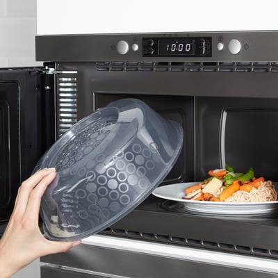 Hand placing clear microwave-safe lid with steam holes over a plate of food in a microwave.
