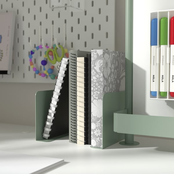 Green desk organiser with bookends holding notebooks and scissors, against white pegboard.