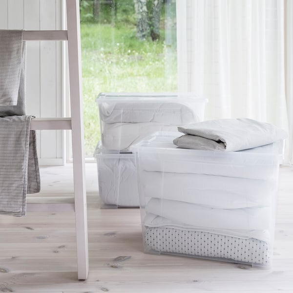 Stacked clear storage boxes with linens, chair, window, bright room.