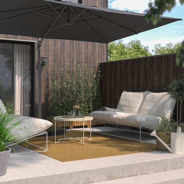 Modern patio with black parasol over white sofa on yellow rug, surrounded by potted plants and a wooden fence with greenery.