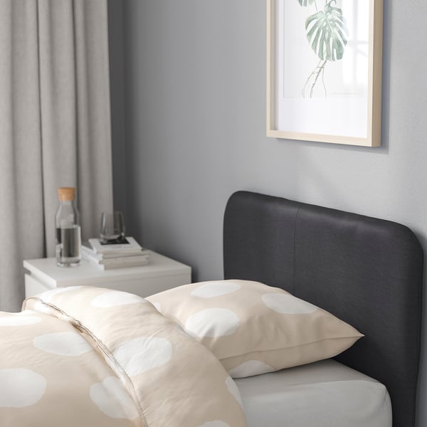 Modern bedroom with black SLATTUM bed, padded headboard, and bedside table with books and carafe. Artwork of green plant hangs above bed.