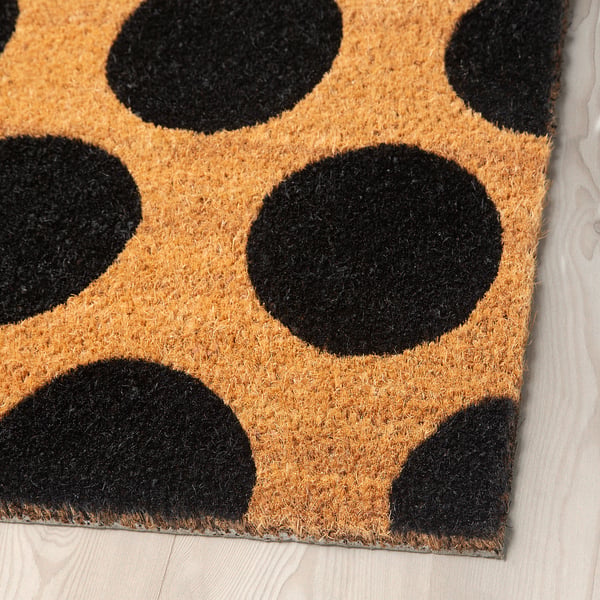 Polka-dotted tan and black doormat with coir fibres, neatly placed on wooden flooring, exuding modern decor.