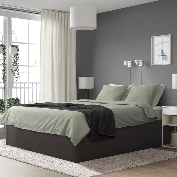 Modern bedroom with TJÅLLE bed, storage, green pillows, dark blanket, tall curtains, small bedside table, lamp, neutral rug.