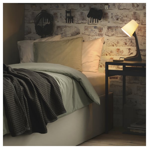 Cosy bed by brick wall, lit lamp, throw pillows, grey blanket, minimalist decor.