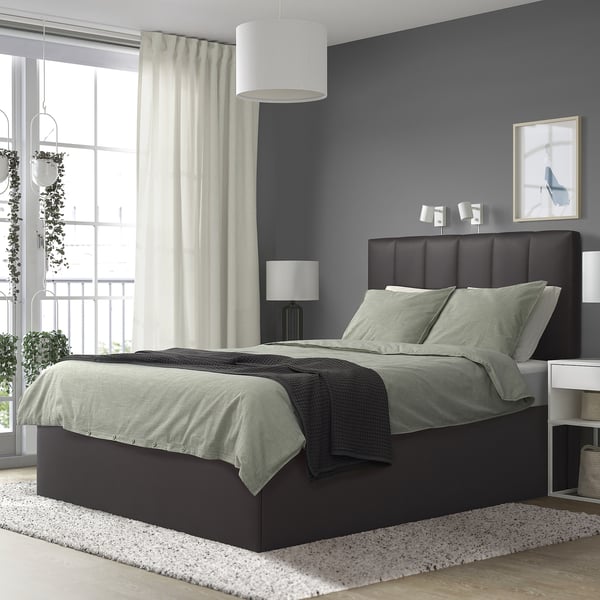Modern dark grey TJÅLLE bed with green linens, large against blue wall. It has a padded headboard and bedside lamps. Rug and balcony seen.