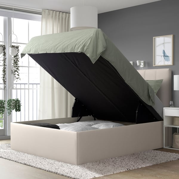 Modern bedroom with TJÅLLE ottoman bed, white headboard, green blanket, and plant. Open storage in bed, bedside table, and soft lighting.