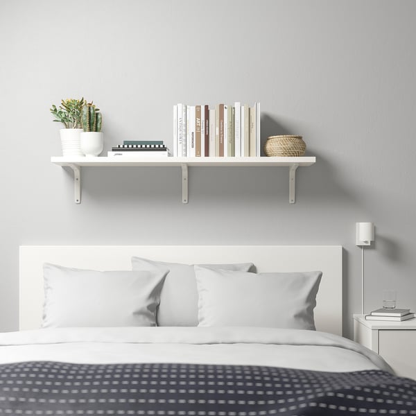 A minimalist bedroom with a TOMTHULT shelf above the bed, holding books, a plant, and a basket. A bedside table with books and a lamp.