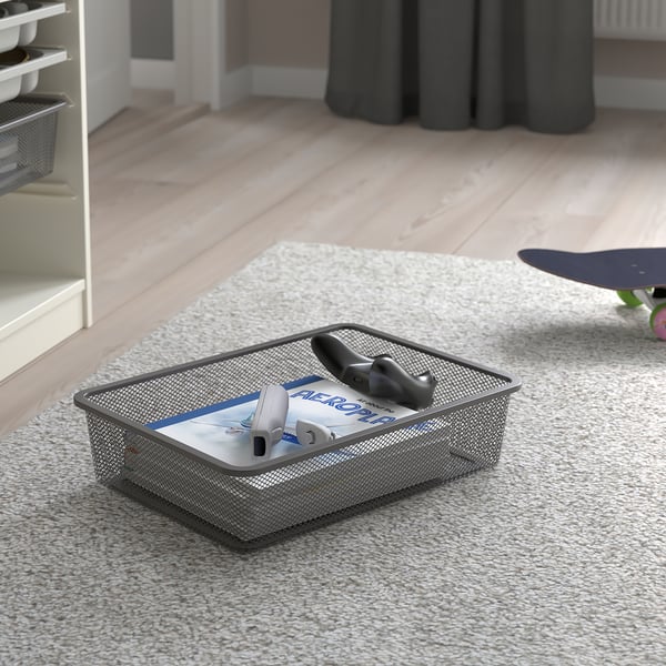 Metal mesh storage box on carpet with gaming controllers and case. TROFAST organisational tool for easy access and tidying.
