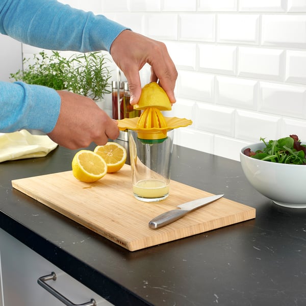 Person squeezing lemon with a bright yellow juicer into a clear glass, with cut lemon halves and knife on a wooden cutting board.