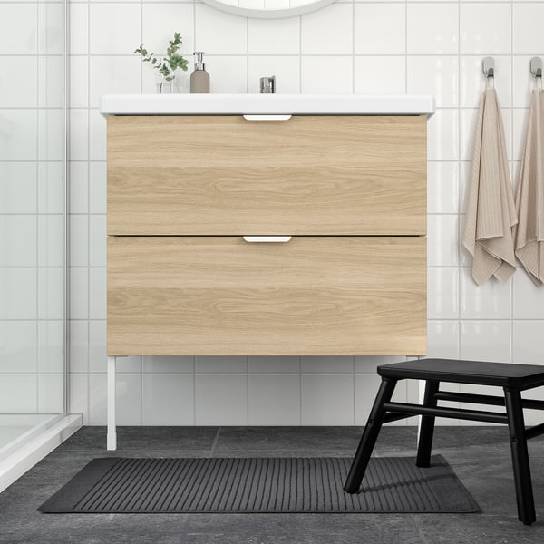 Modern bathroom with light wood vanity, black stool, grey mat, white walls & glass door.