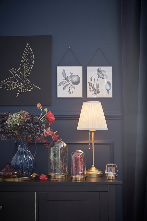 Dark blue wall with art, lamp, flowers, and decor. Golden ÅRSTID lamp has fabric shade, brass base.